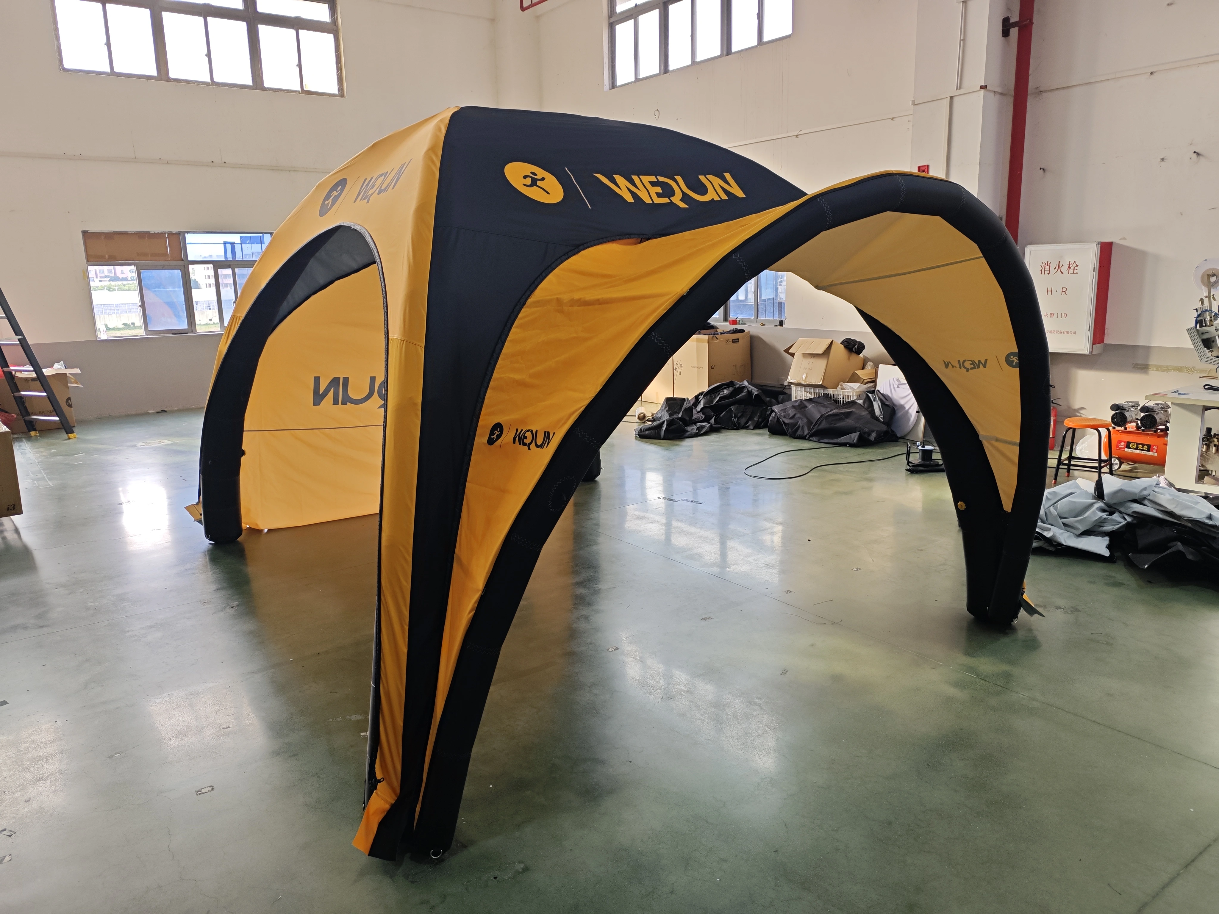 Another angle of the yellow and black "WEQUN" inflatable arch tent, positioned on the green floor of a manufacturing facility, with cardboard boxes and factory equipment visible in the background.